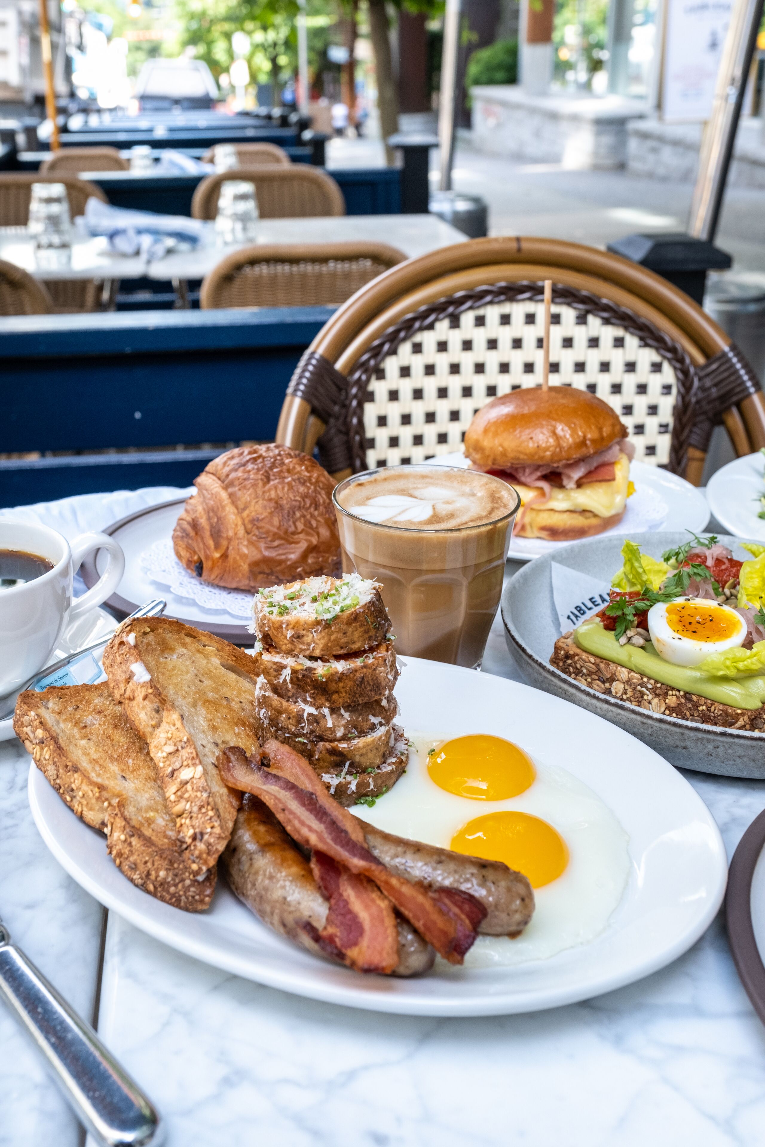 A breakfast spread on a table with toast, eggs, bacon, a croissant, a sandwich, and a drink.