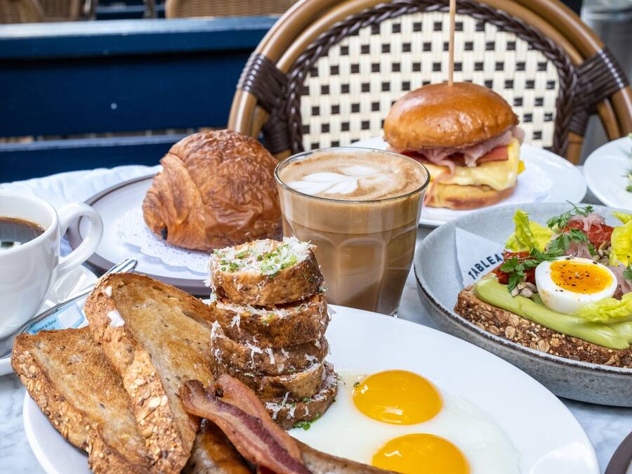A breakfast spread on a table with toast, eggs, bacon, a croissant, a sandwich, and a drink.
