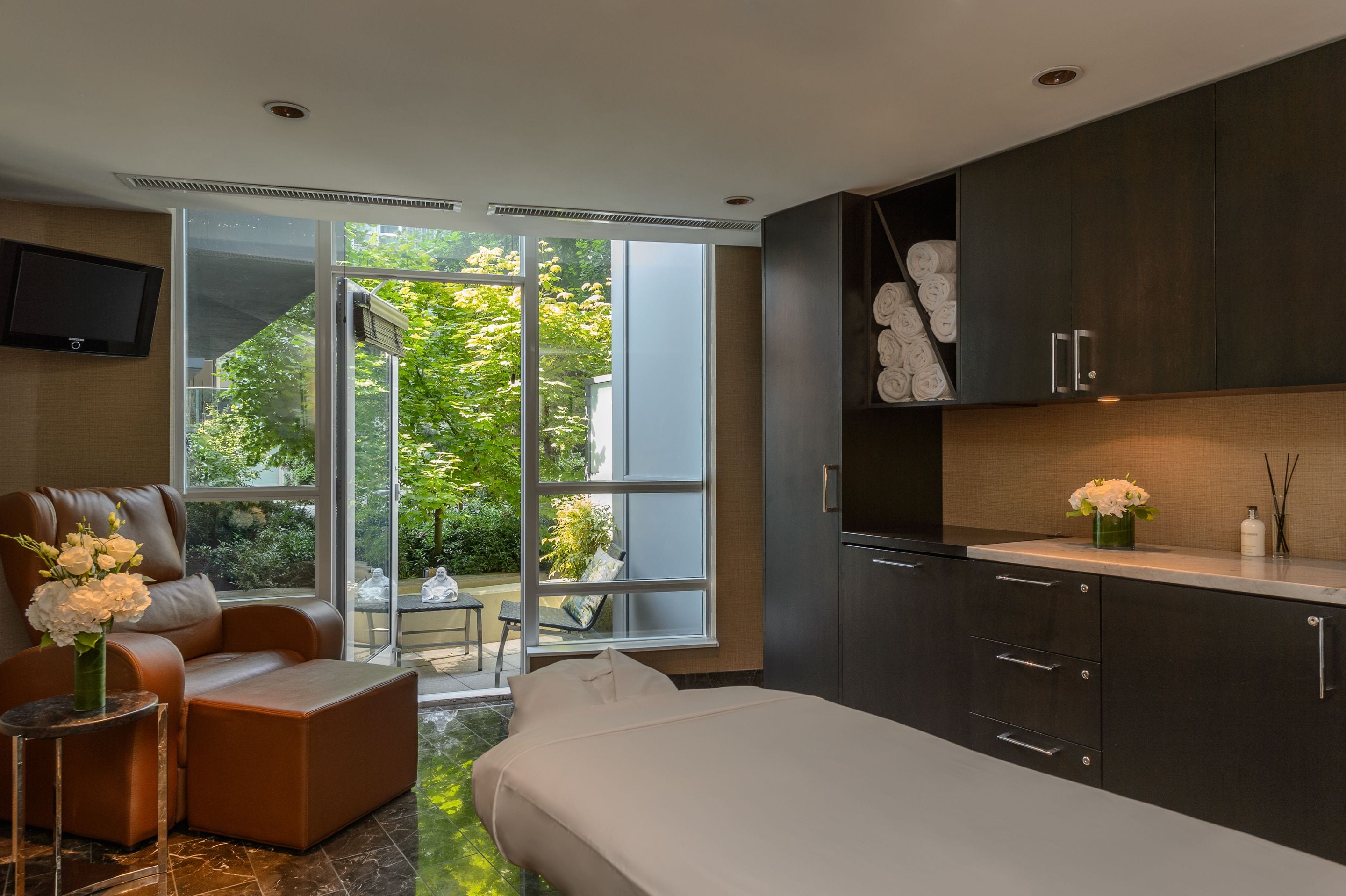 A modern hotel room with a large sliding glass door to a garden, a sofa, a bed, and dark cabinetry.
