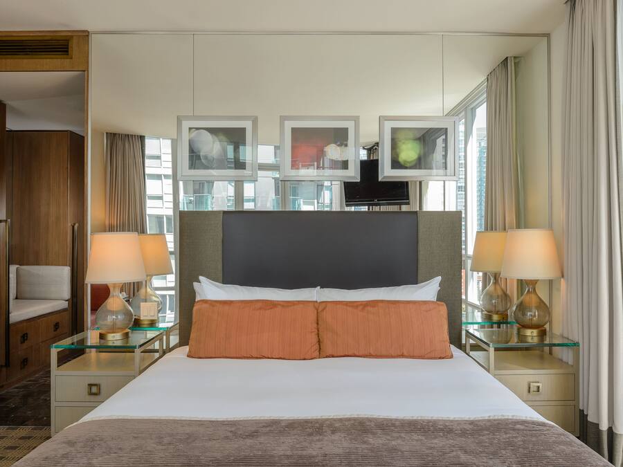 A hotel room with a large bed, two bedside tables with lamps, a headboard with framed pictures, and a view of the city through large windows.