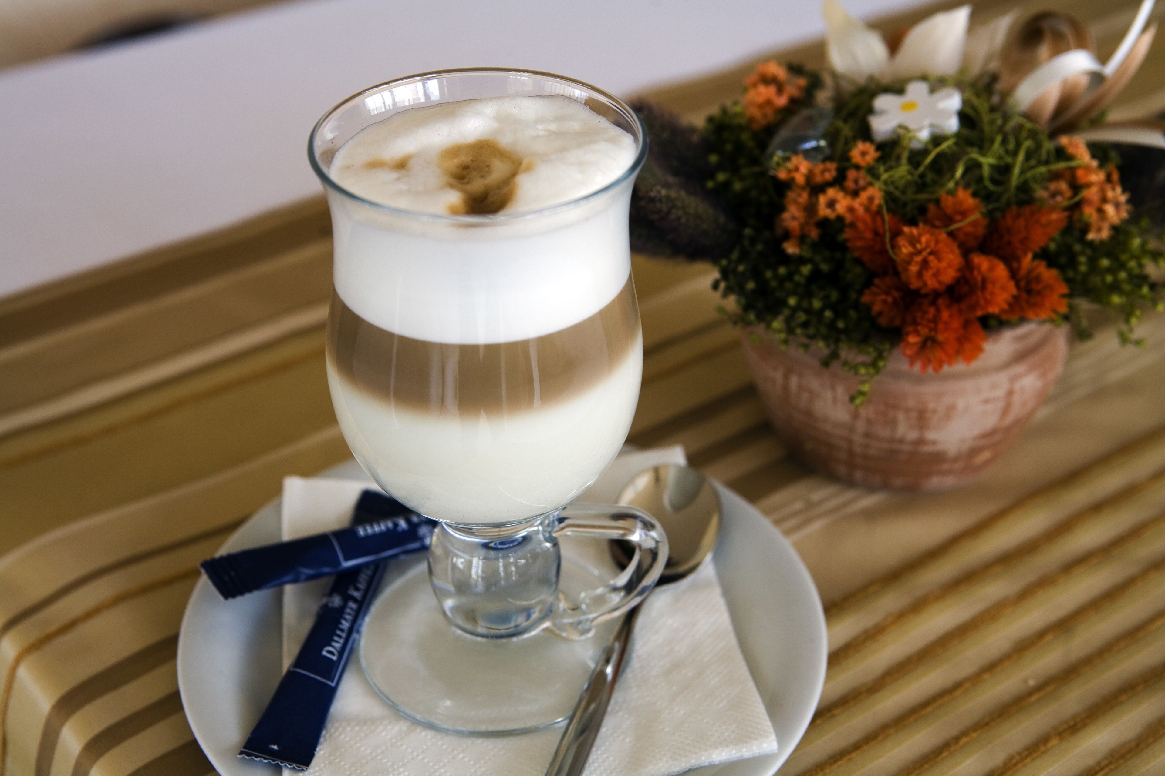 Ett glas med en dryck i flera lager, placerat på en liten tallrik tillsammans med en sked och en servett, en dekorativ blomsteruppsättning och ett randigt bordsduk.