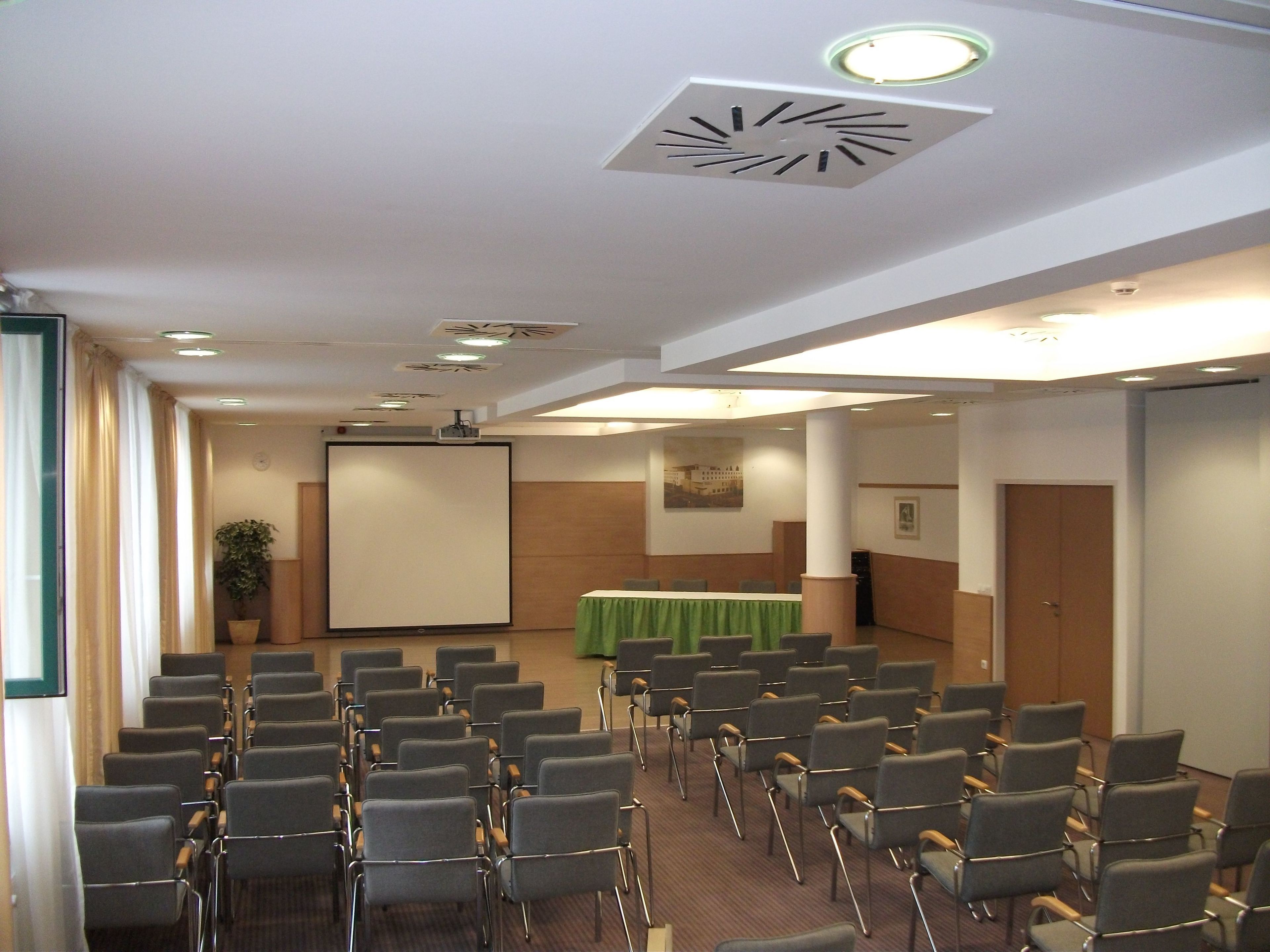 A conference room with rows of chairs, a projector screen, a table with a green tablecloth, and a clock on the ceiling.