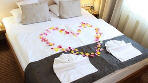 A neatly made bed with a heart-shaped arrangement of rose petals and two folded towels on a dark grey bedspread.