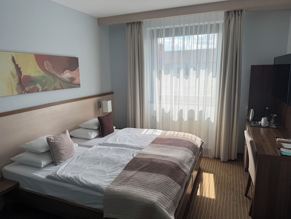 Habitación de hotel con una cama grande, un escritorio, un televisor y una ventana con cortinas traslúcidas.