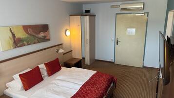 A hotel room with a bed, a nightstand, a lamp, a painting on the wall, an air conditioning unit, and a door.