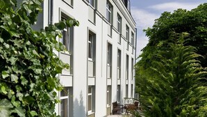 Un bâtiment moderne à la façade symétrique, doté de grandes fenêtres et agrémenté d’un jardin bien entretenu, où l’on trouve diverses plantes et un espace aménagé pour s’asseoir.