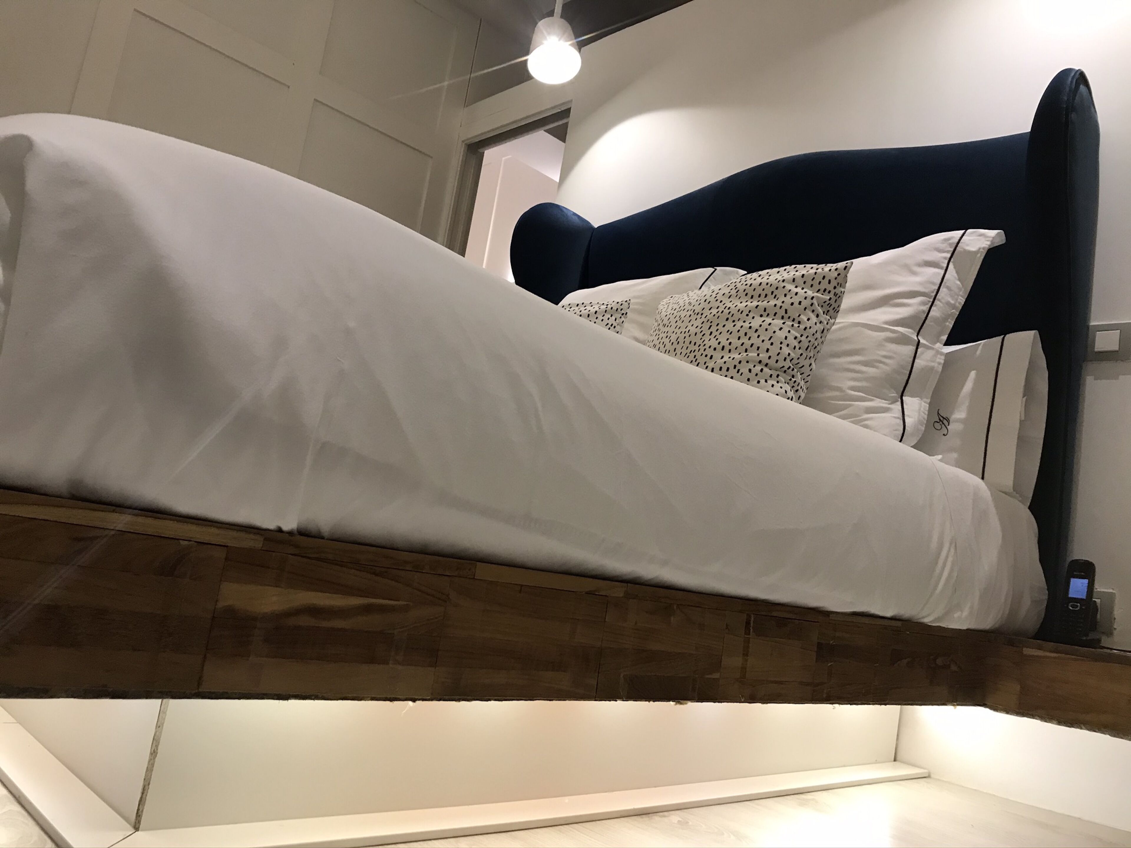 Una cama con cabecera oscura y ropa de cama blanca, suelo de madera y una lámpara colgante.
