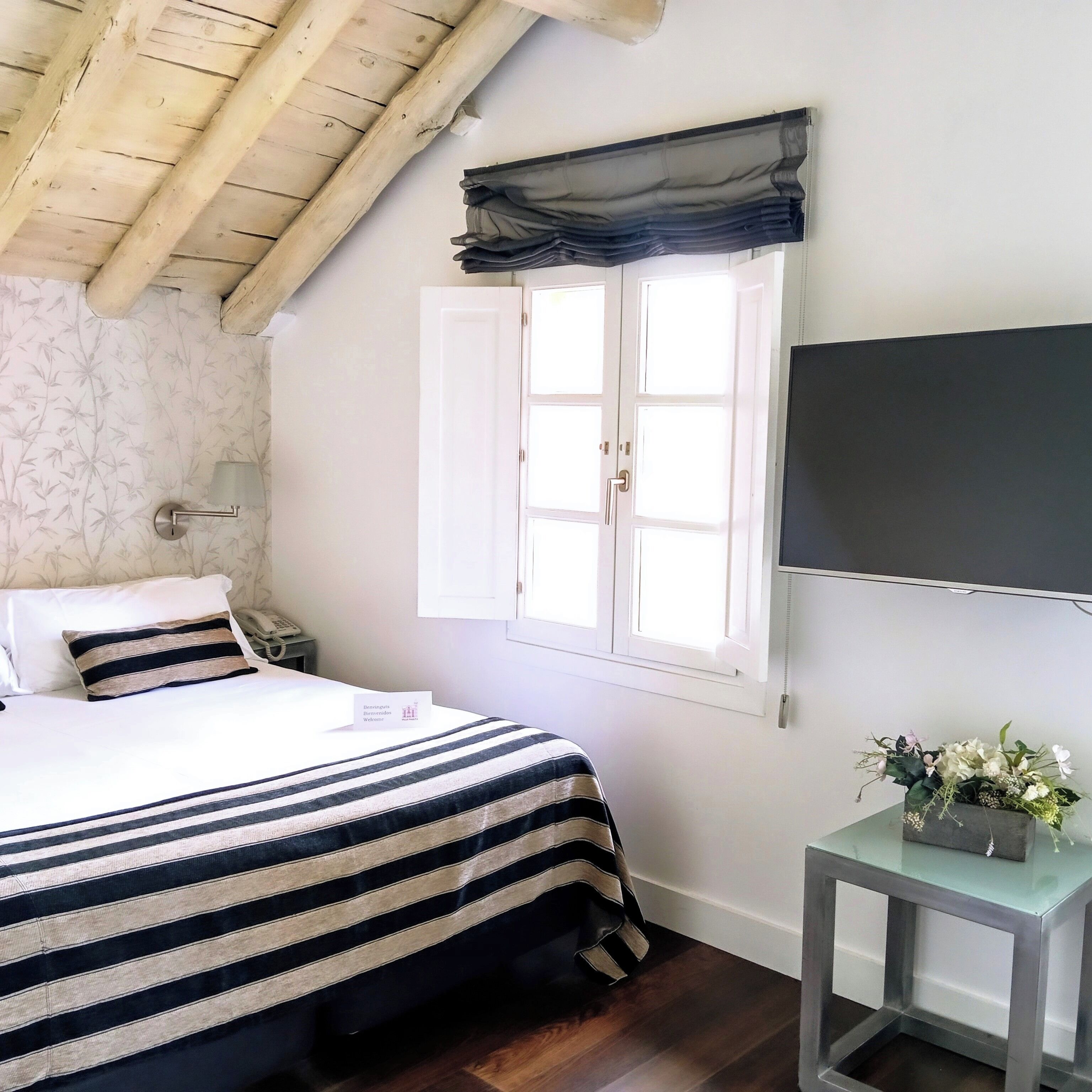 Un dormitorio con cama, una mesita de noche con un florero, una ventana con cortinas y un televisor en la pared.