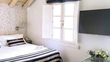 Un dormitorio con cama, una mesita de noche con un florero, una ventana con cortinas y un televisor en la pared.