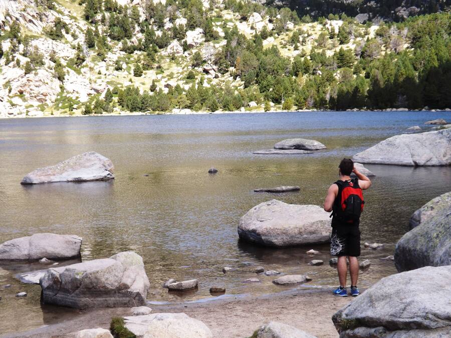 Una persona de pie junto a un lago con rocas grandes y árboles al fondo.