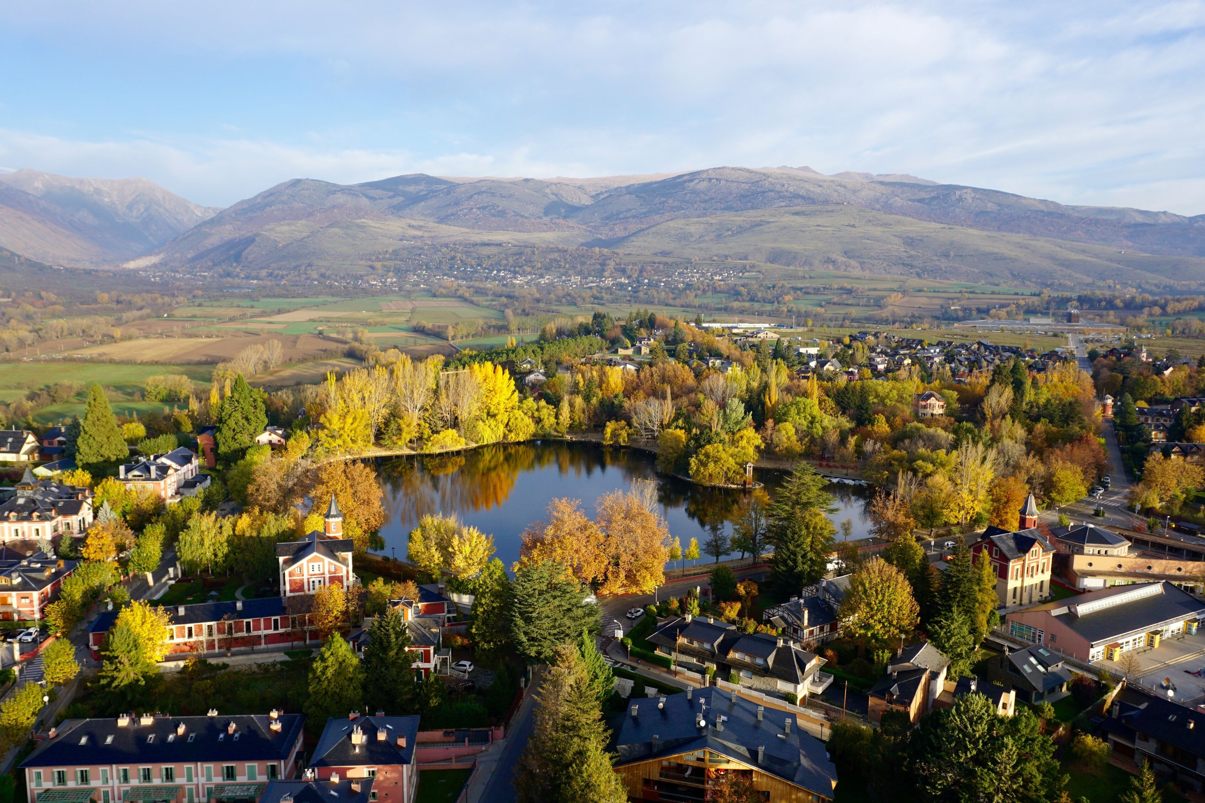 Un pueblo con un lago rodeado de árboles de colores otoñales y casas, con montañas al fondo.