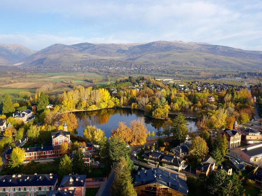 Un pueblo con un lago rodeado de árboles de colores otoñales y casas, con montañas al fondo.