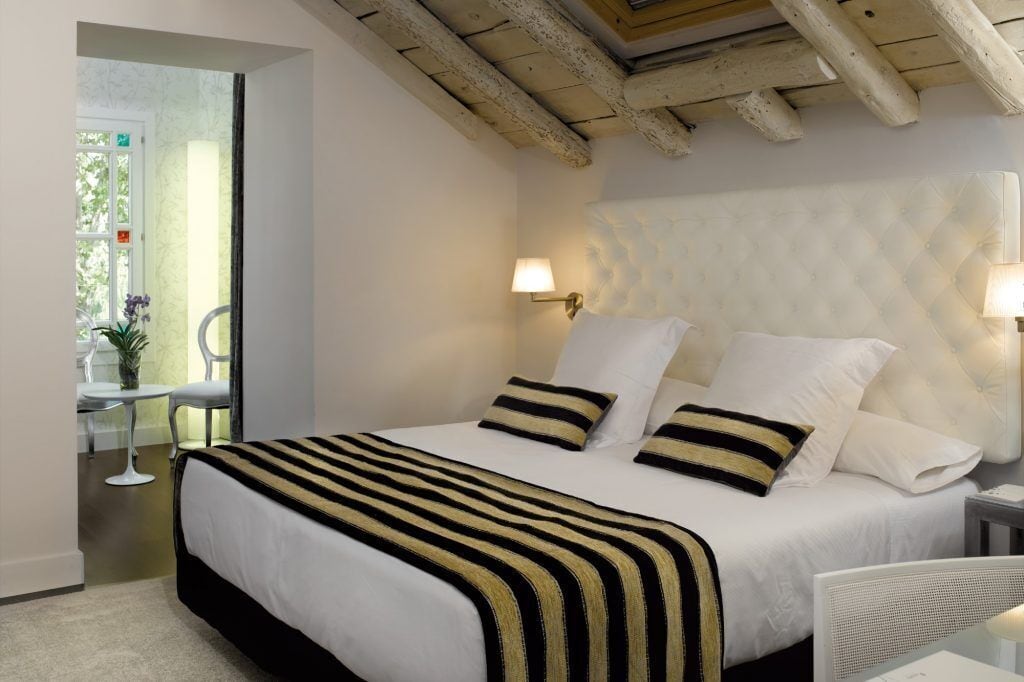 Habitación de hotel con cama, lámparas de noche, una mesita con silla y vistas al exterior.