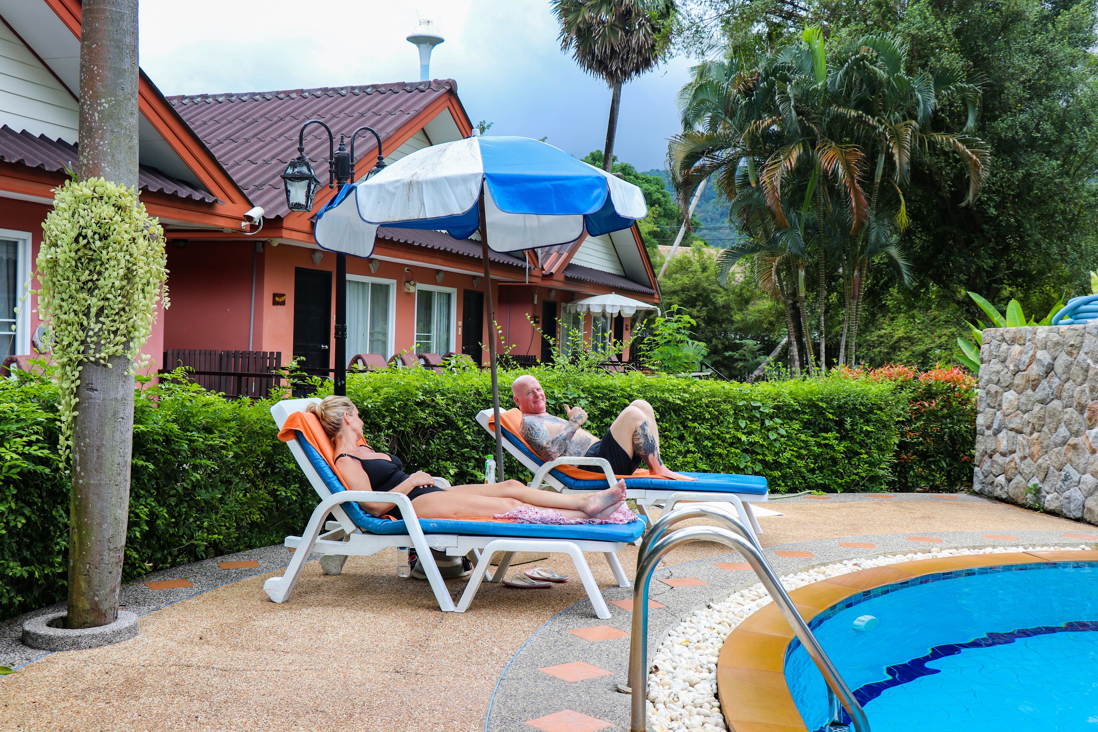 Zona junto a la piscina con sillones de descanso y una sombrilla grande, rodeada de vegetación y un edificio con techo de tejas.