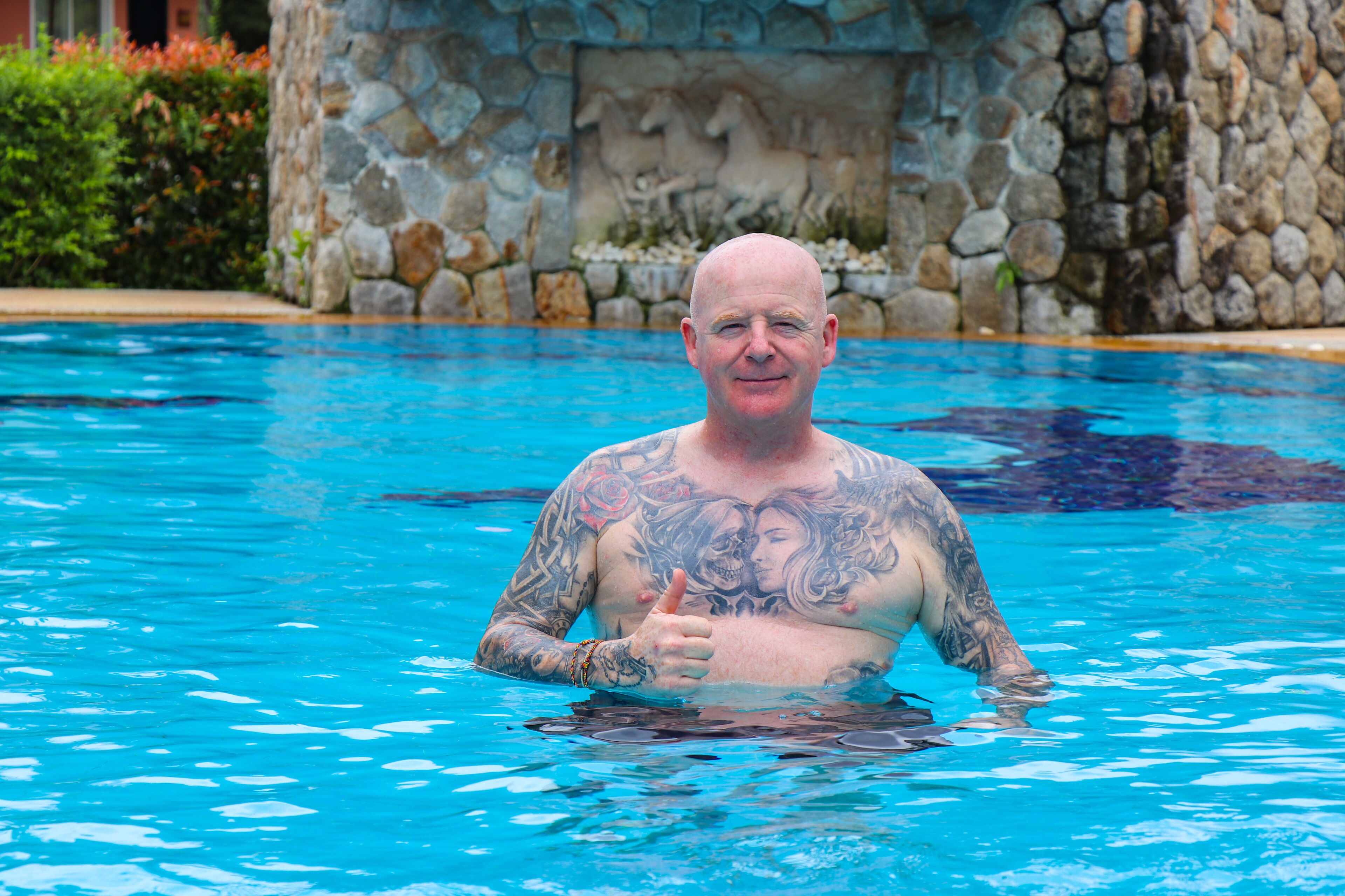 Un homme dans une piscine, portant des tatouages, entouré d’un mur de pierre et de verdure.