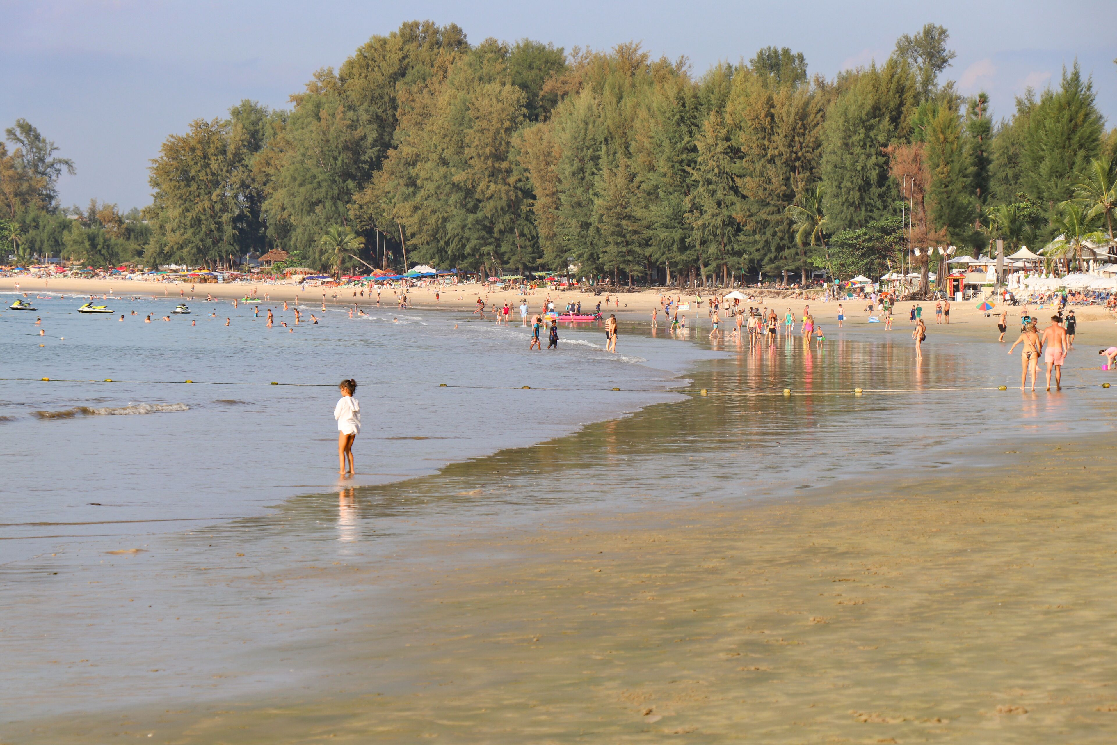 Una playa con agua cristalina, gente nadando y un área boscosa al fondo.