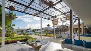 A spacious outdoor lounge area with white wicker furniture, a pool, and a clear view of the resort's buildings.