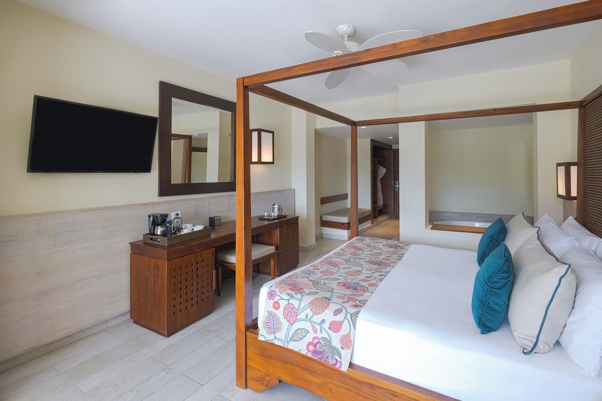 A four-poster bed with a floral bedspread, a wooden desk with a coffee maker, a flat-screen TV mounted on the wall, and a mirror above the desk.