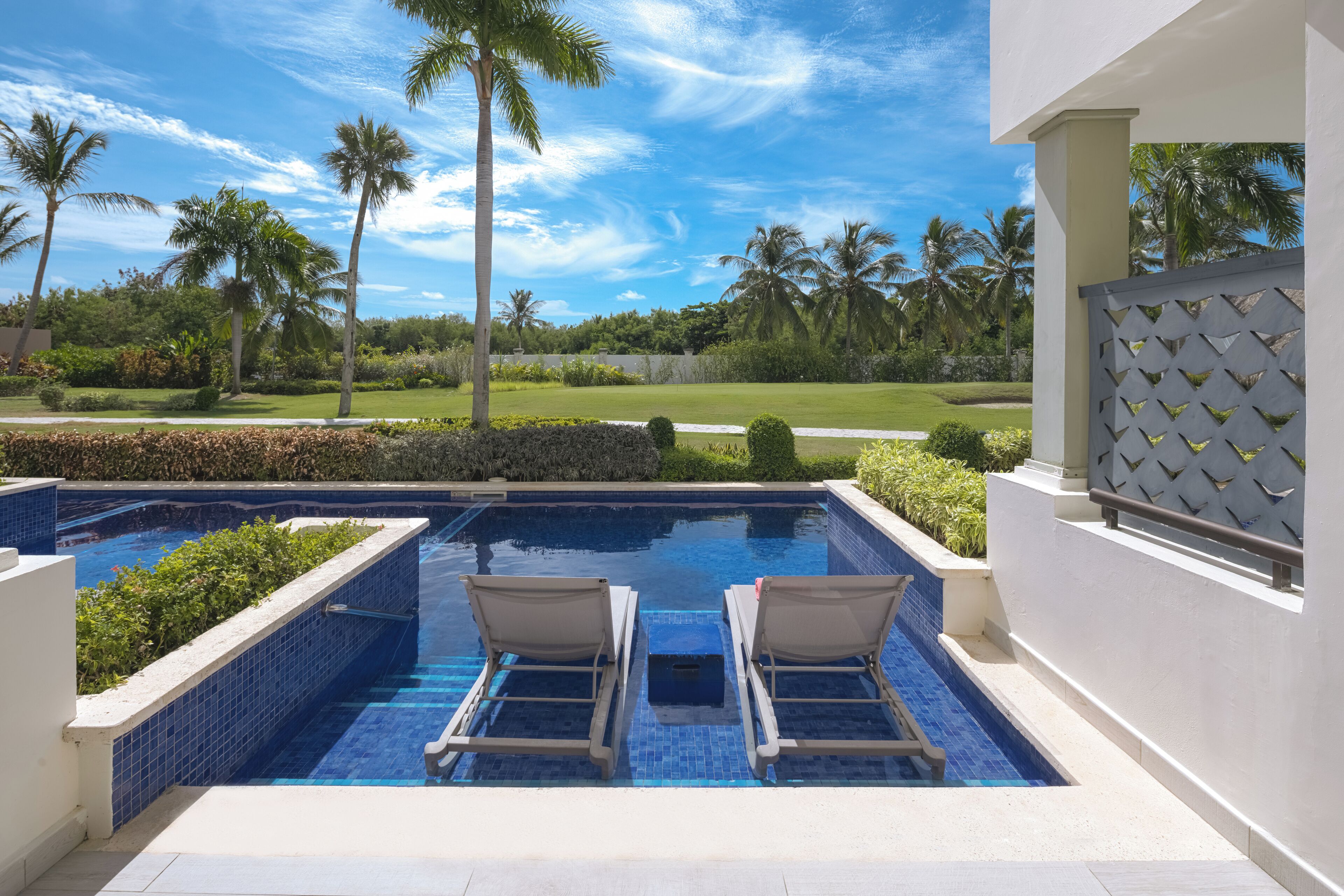 A rectangular swimming pool with two lounge chairs facing the water, surrounded by a white wall and a decorative fence.
