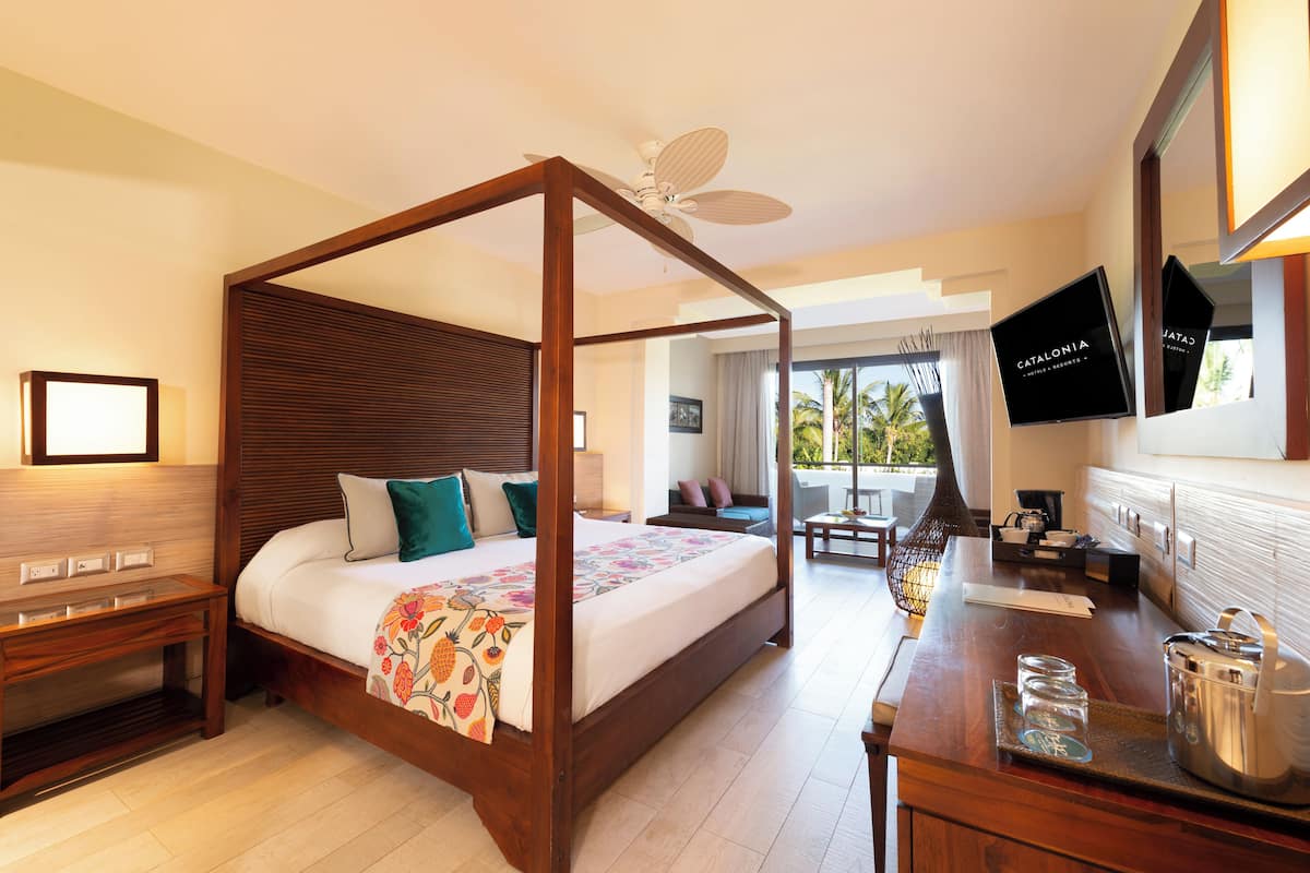 A spacious hotel room with a four-poster bed, a balcony with a view of palm trees, and a flat-screen TV mounted on the wall.