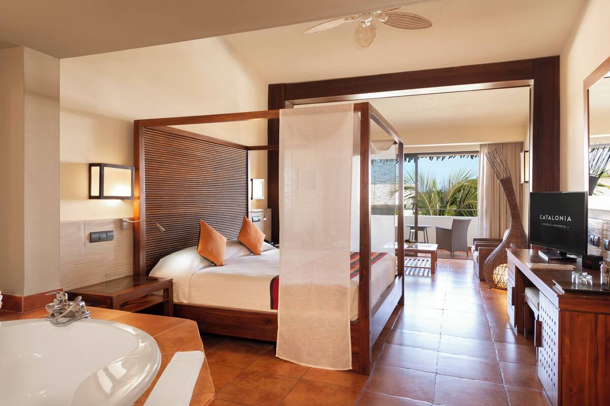 A hotel room with a large bed, a separate bathroom with a bathtub, a TV, and a balcony with a view of palm trees.