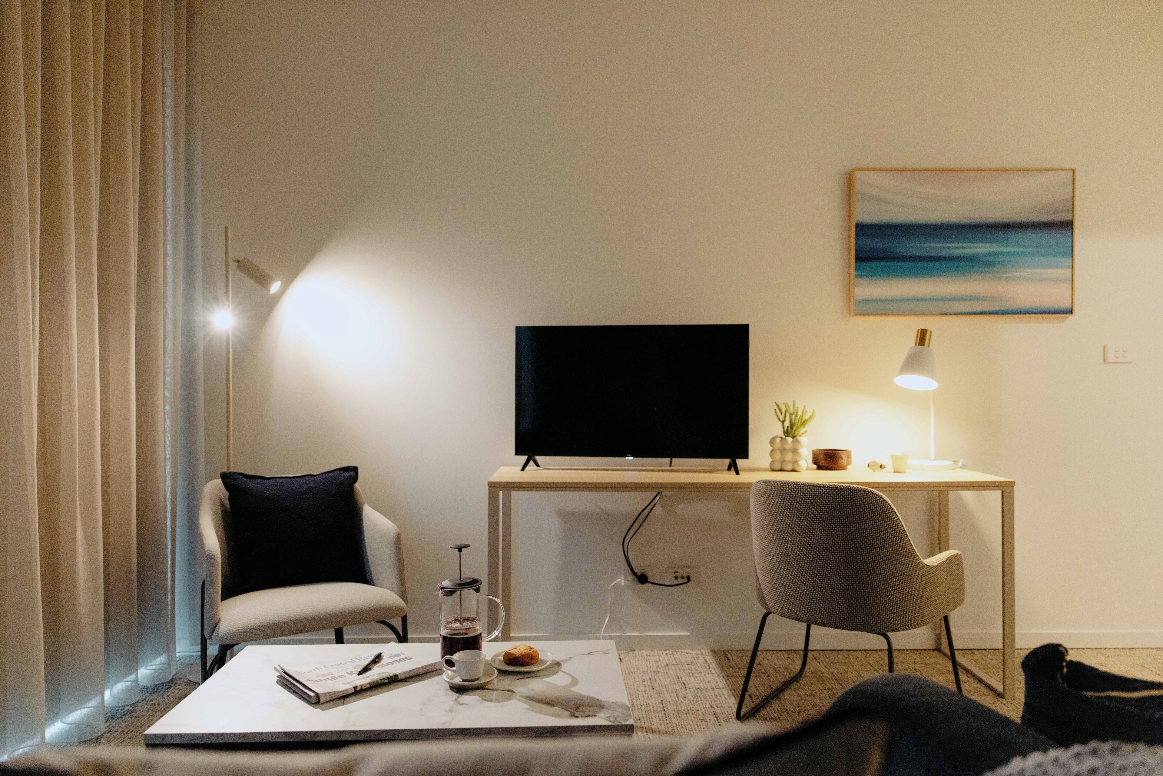 Une chambre d’hôtel moderne équipée d’un téléviseur à écran plat, d’un bureau avec une lampe, d’une chaise, d’une table basse avec un livre et une cafetière, ainsi que d’un tableau au mur.