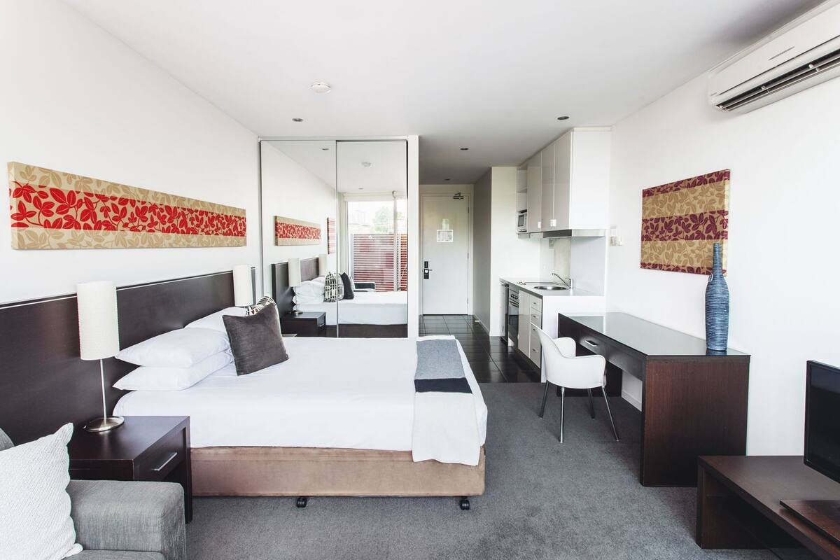 A hotel room with a bed, bedside tables, a desk, a chair, a mirror, and a kitchenette.