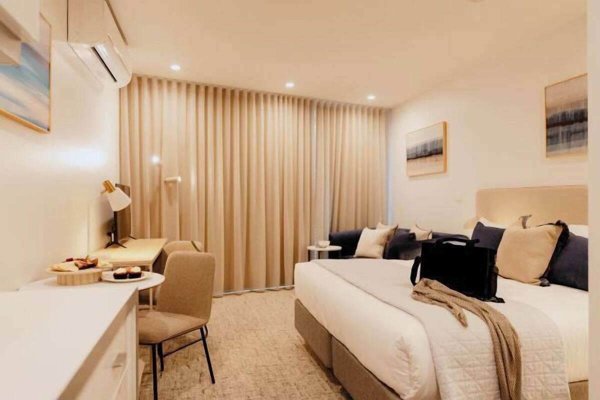 A hotel room with a bed, a desk, a chair, a lamp, and a large window with curtains.