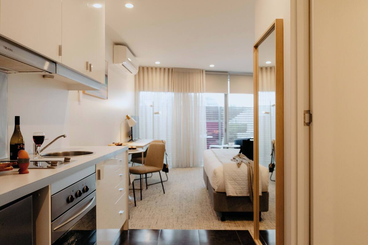 A compact hotel room with a kitchenette, a bed, a desk, and a large window.