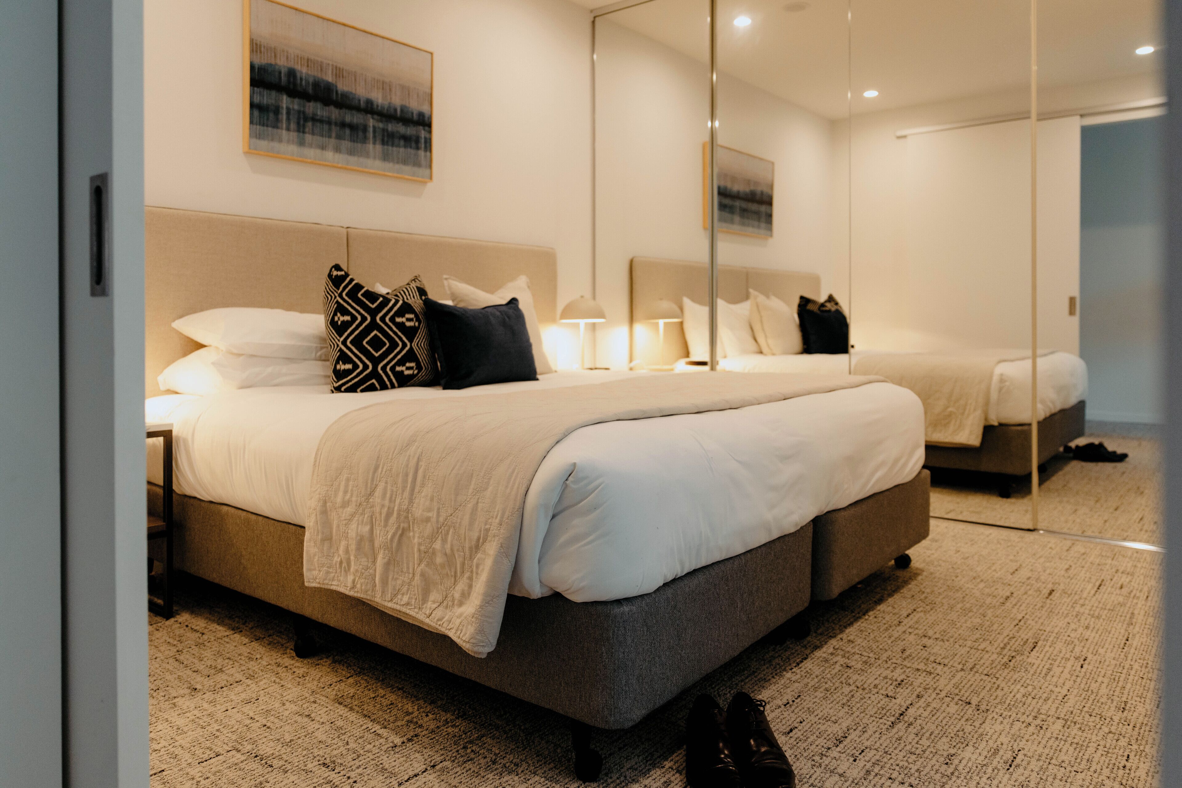 A modern hotel room with a large bed, a bedside table, a mirror, and a carpeted floor.