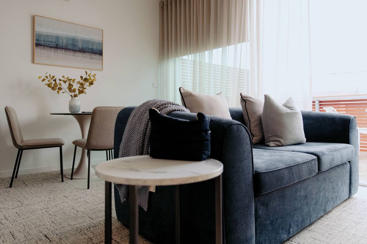 A modern living room with a dark blue sofa, a round coffee table, and a small side table with a vase of dried flowers.