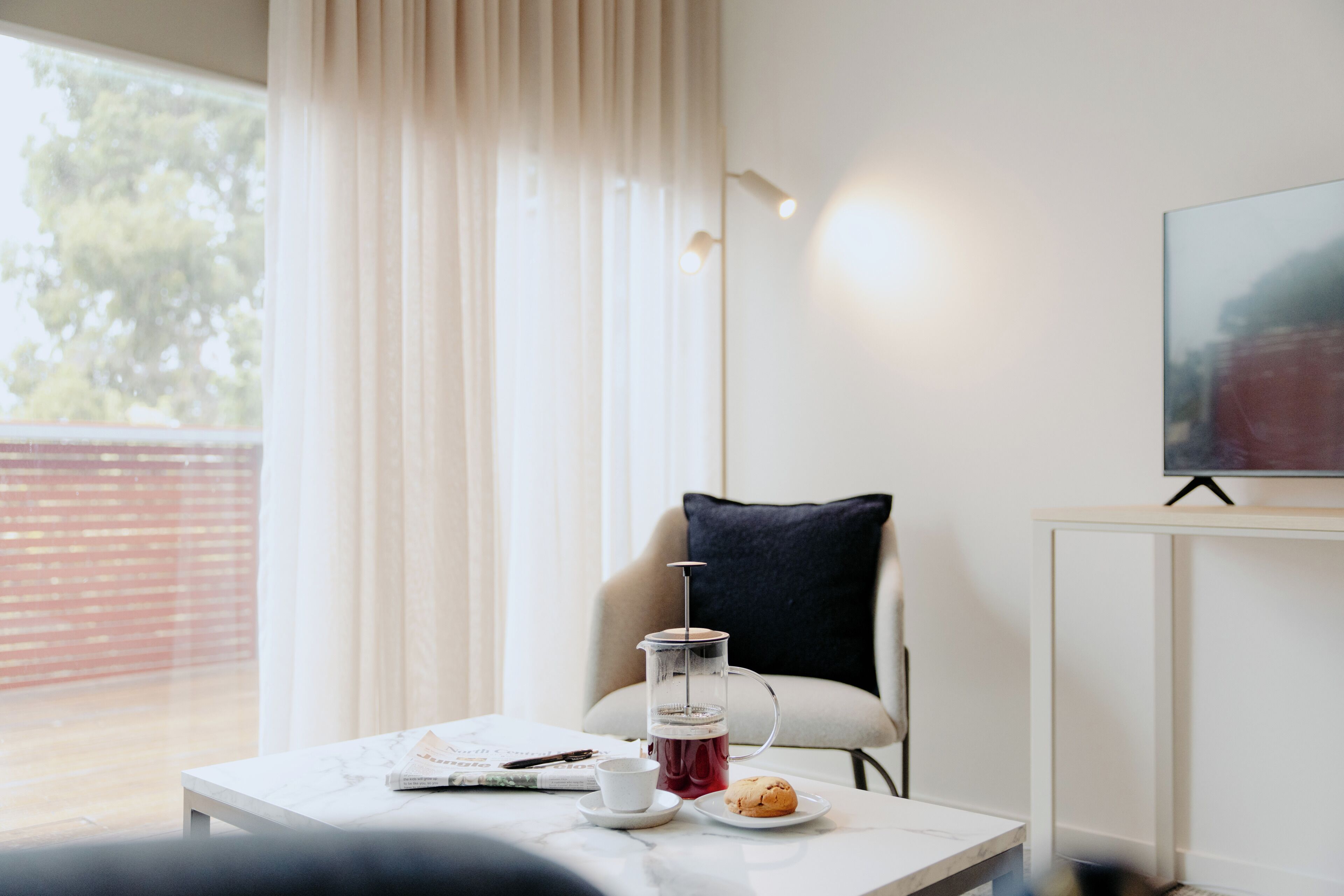 A modern room with a coffee table, a glass carafe, a cup, a plate with a pastry, a chair, a TV, and a window with curtains.