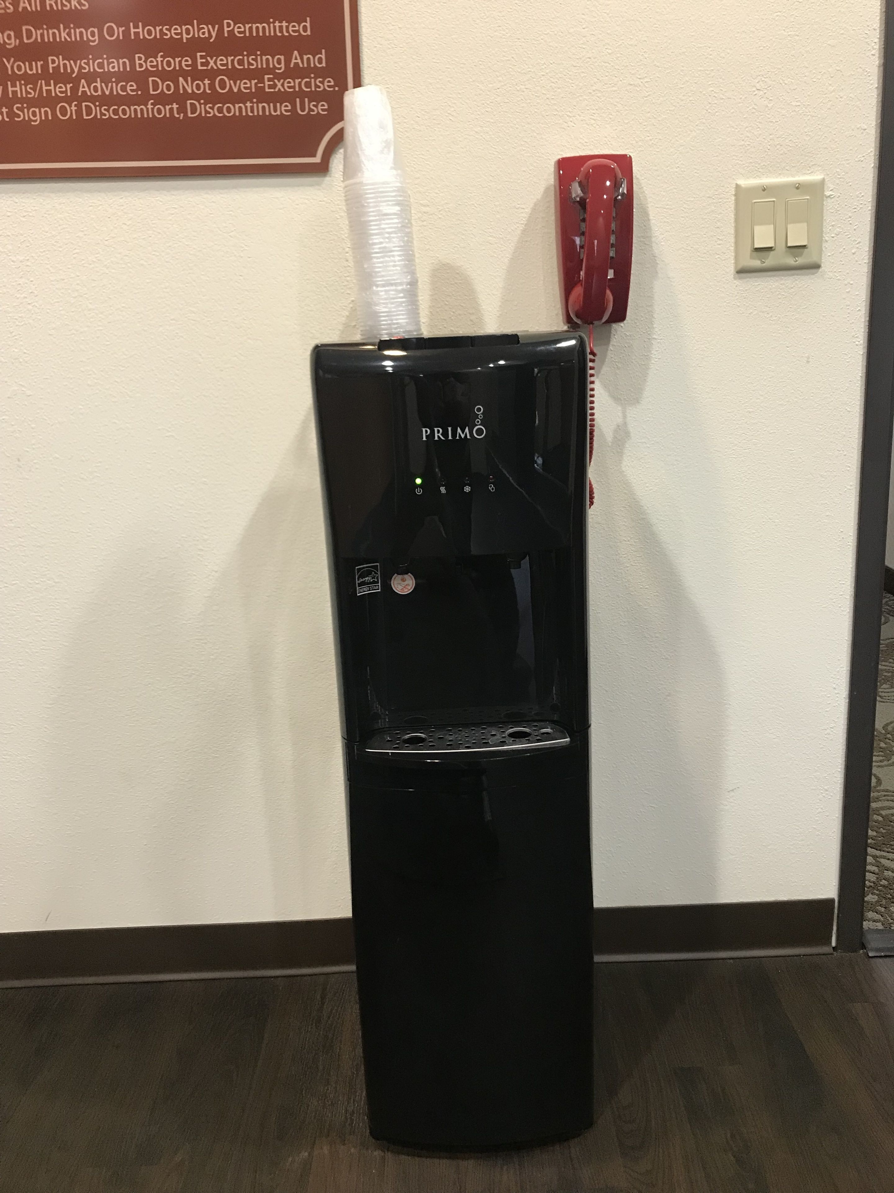Un dispensador de agua negro con la marca PRIMO, un teléfono instalado en la pared y un letrero con consejos sobre salud y ejercicio.