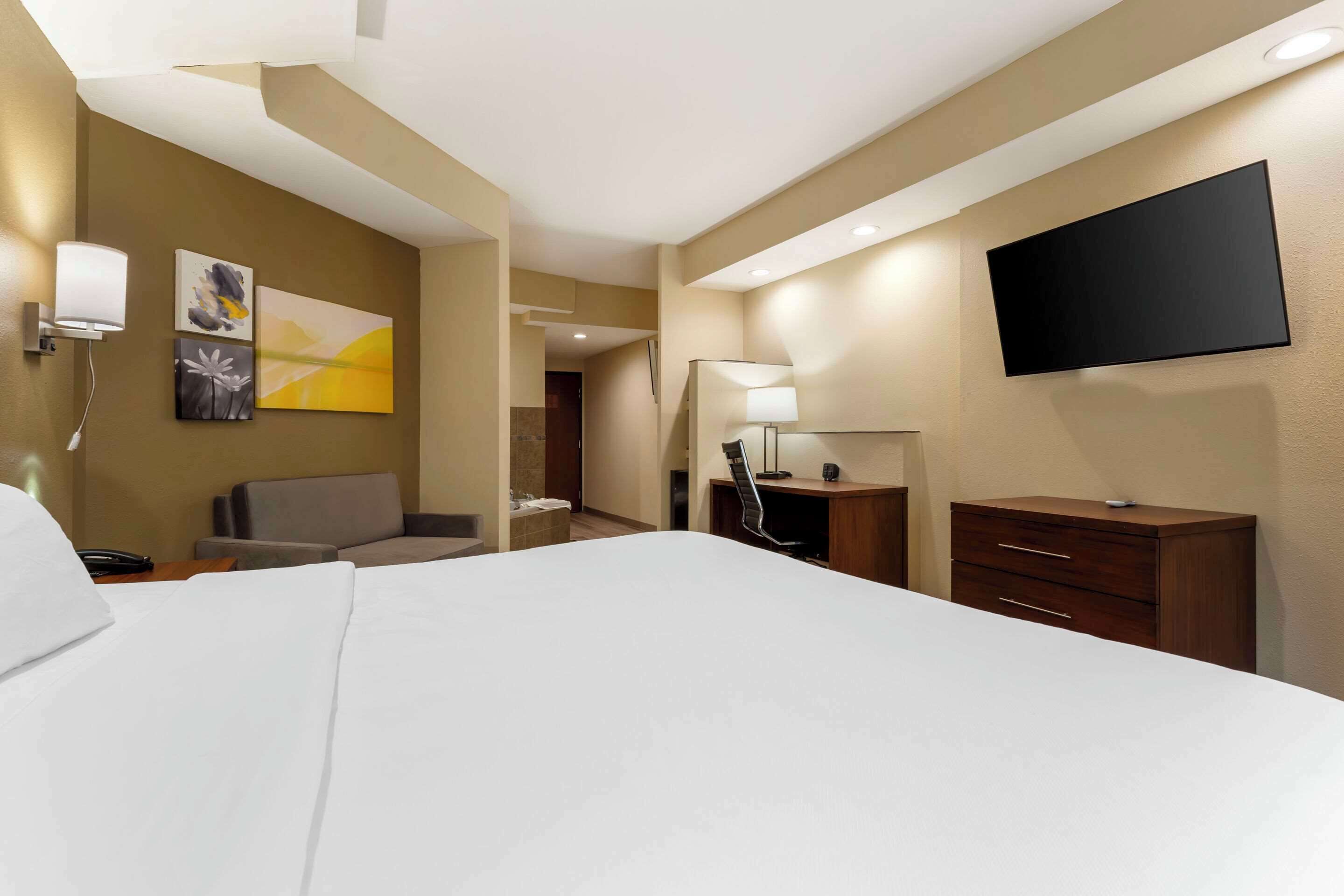 A hotel room with a large bed, a flat-screen TV mounted on the wall, a desk with a lamp, and a chair.
