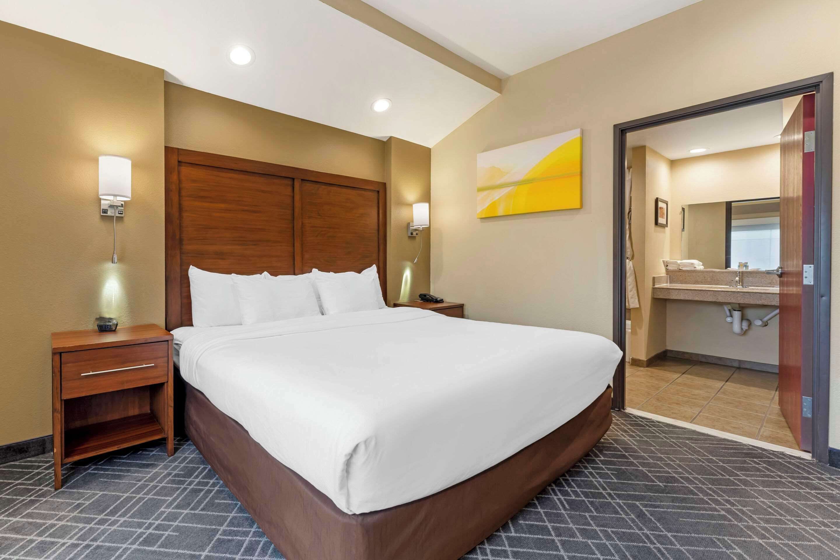 Habitación de hotel con una cama grande, mesitas de noche, una lámpara de pared y un baño visible a través de una puerta abierta.