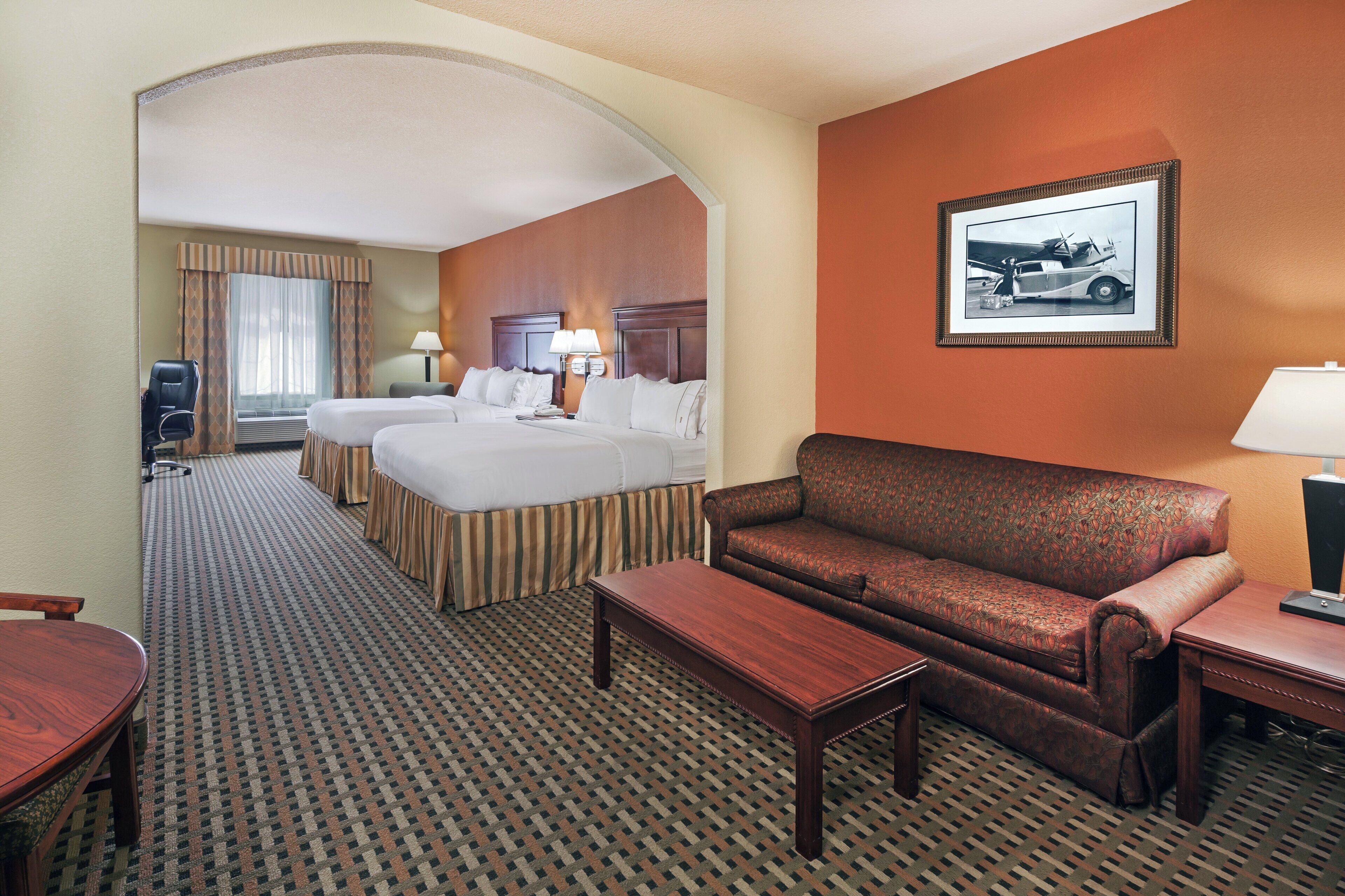 A hotel room with two beds, a sofa, a small table, a desk, a lamp, and a framed picture on the wall.