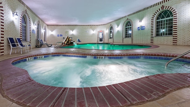 En indendørs swimmingpool med murstensvægge og buede vinduer, omgivet af liggestole og en fliseterrasse.