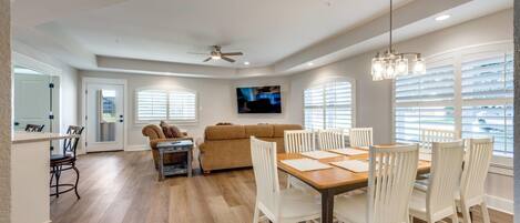 A spacious living area with a dining table, white chairs, a sofa, and a TV mounted on the wall.
