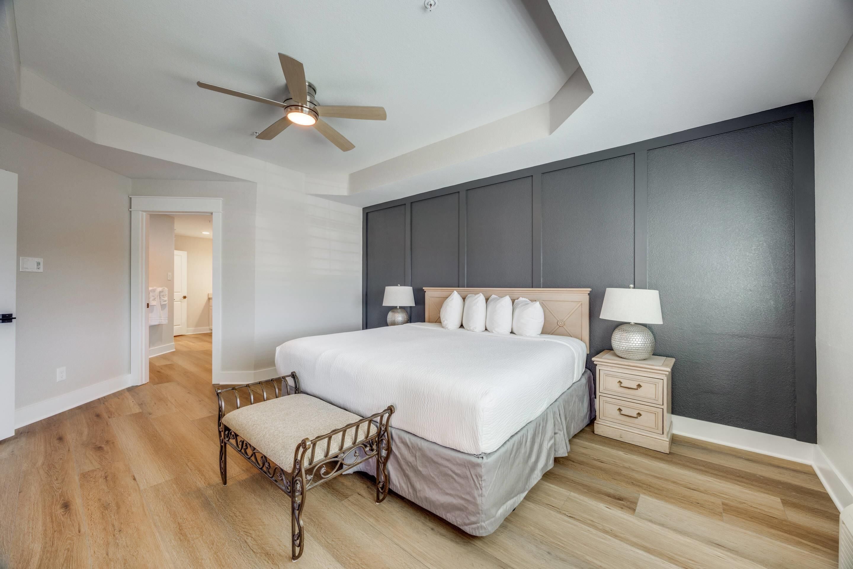 A bedroom with a large bed, a wooden bench, a nightstand, a ceiling fan, and a view of a hallway.