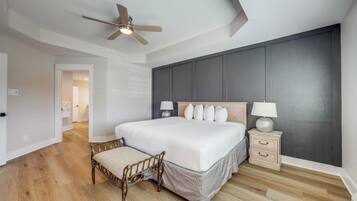 A bedroom with a large bed, a wooden bench, a nightstand, a ceiling fan, and a view of a hallway.