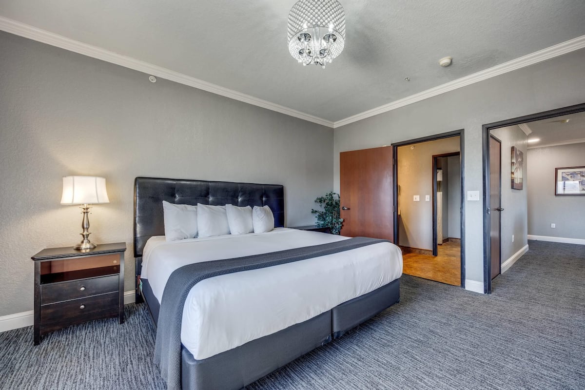 Ein Hotelzimmer mit einem großen Bett, einem Nachttisch mit Lampe, einer Kristallleuchtre und einer Tür zu einem anderen Raum.