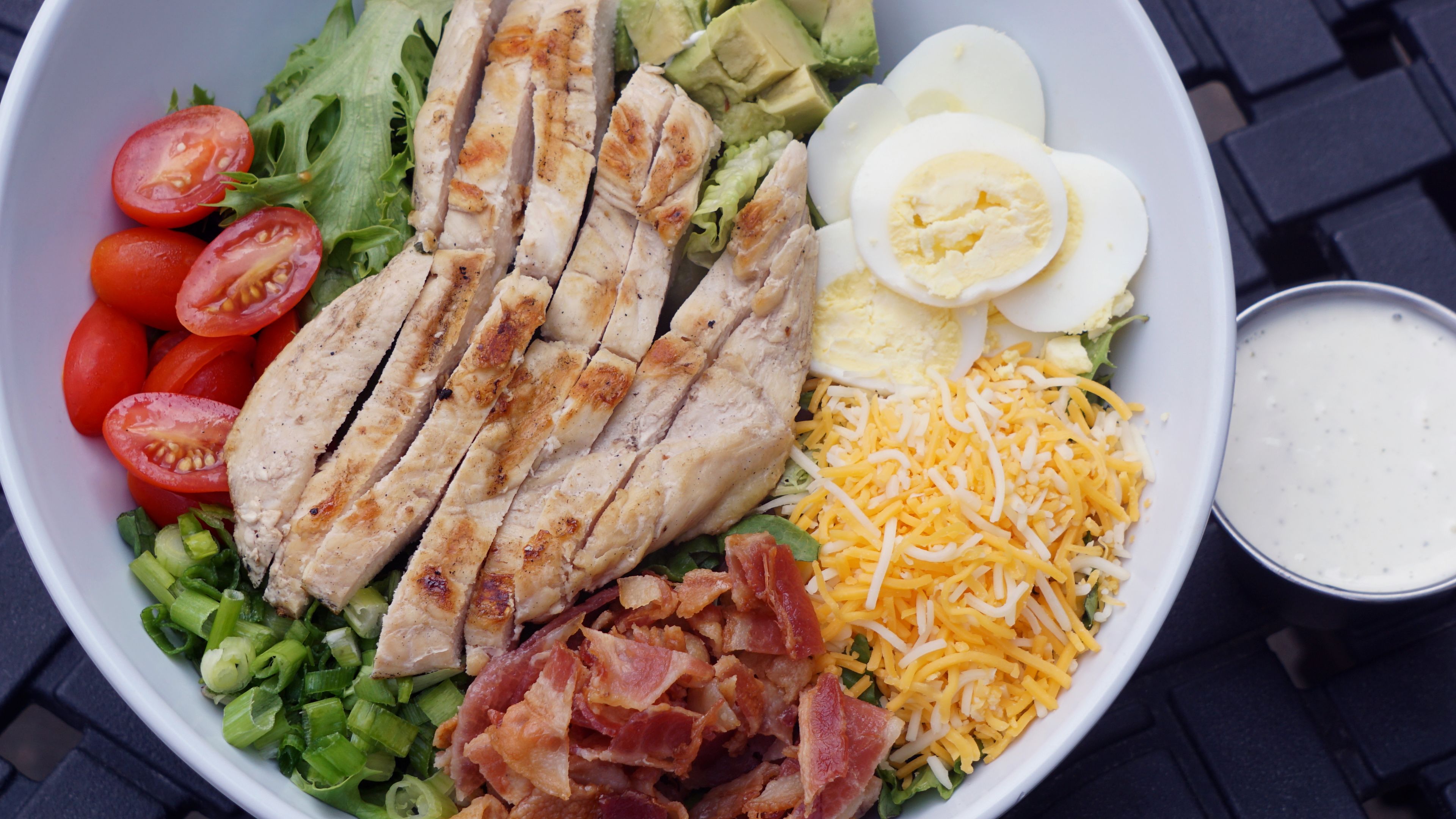 A salad bowl with various ingredients including lettuce, tomatoes, chicken, avocado, boiled eggs, cheese, and bacon.