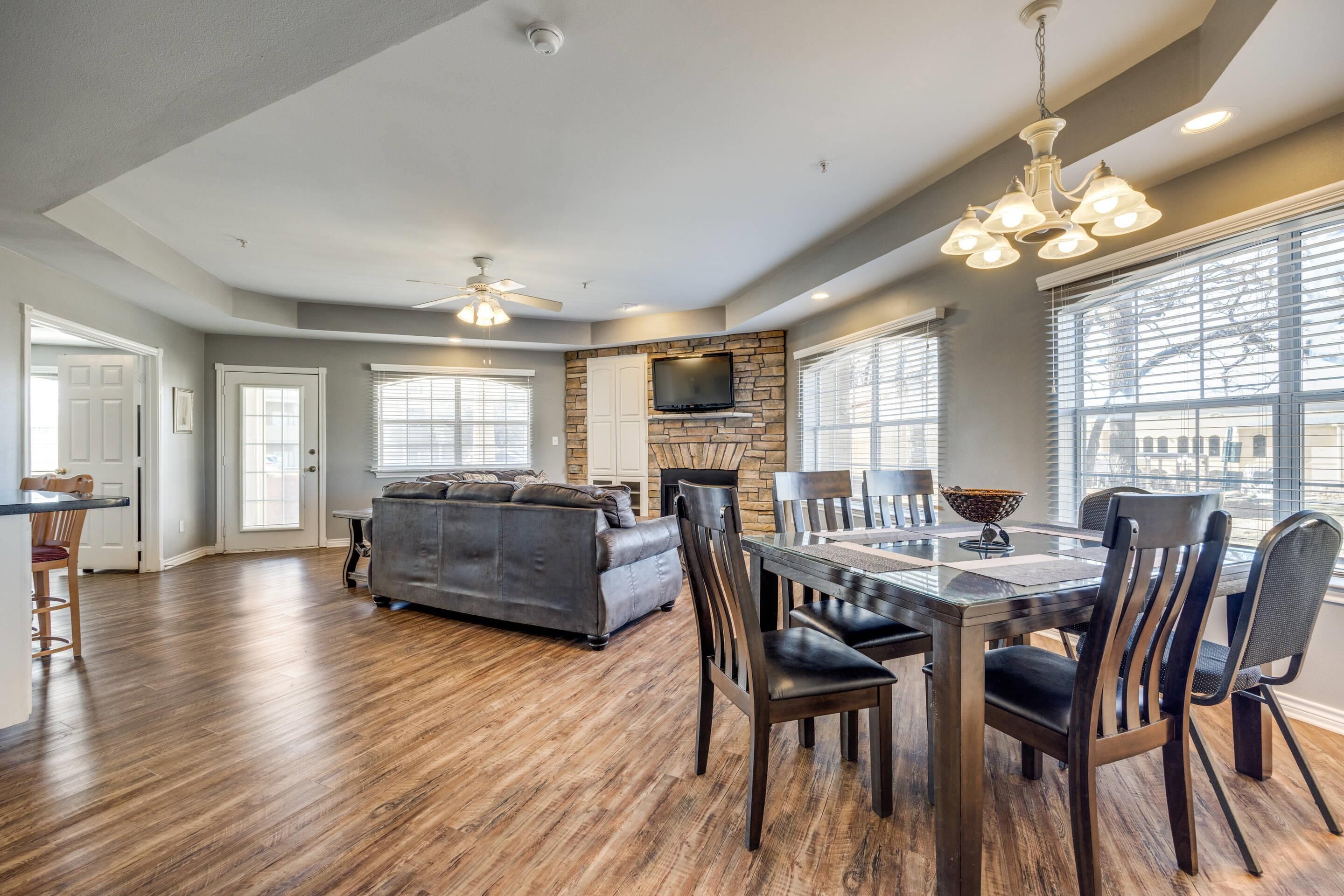 A spacious living room with a gray sofa, wooden dining table, and chairs. A stone fireplace is visible, and there are large windows with blinds.