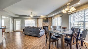 A spacious living room with a gray sofa, wooden dining table, and chairs. A stone fireplace is visible, and there are large windows with blinds.
