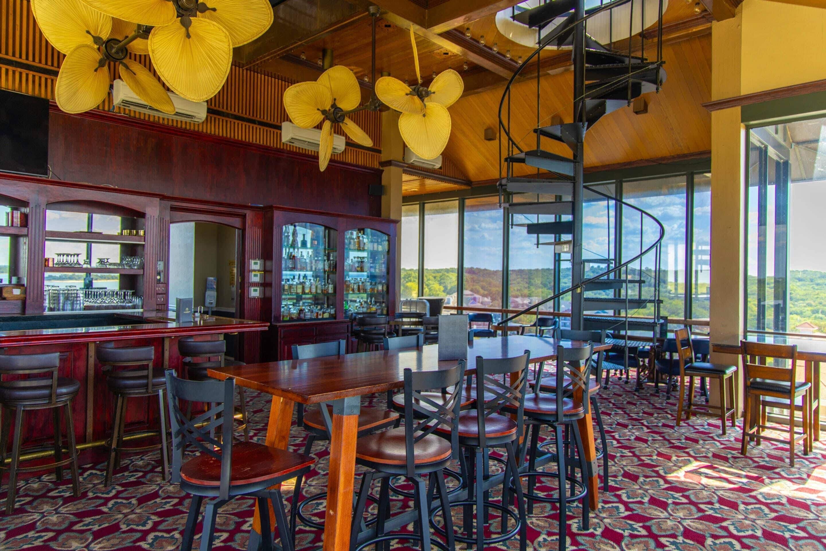 A bar area with a spiral staircase, wooden tables, and chairs, and a view of the landscape.