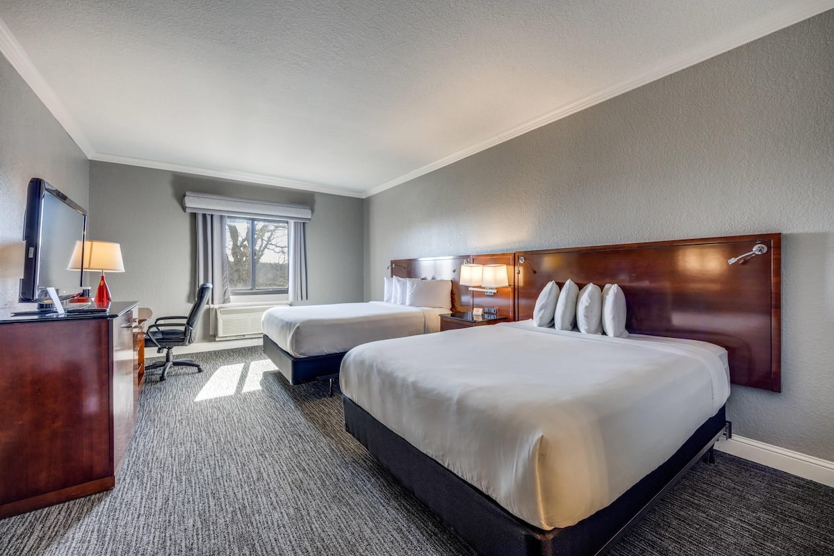 A hotel room with two beds, a desk, a chair, a lamp, and a window.