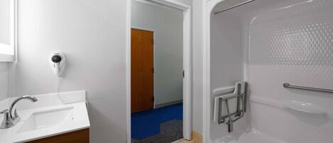Un baño con un lavamanos blanco, una ducha con barra de apoyo y una puerta que da a otra habitación.