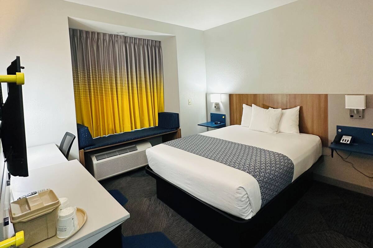 A hotel room with a large bed, a desk with a chair, a small table, and a chair. There is a window with curtains, a wall-mounted phone, and a wall-mounted lamp.