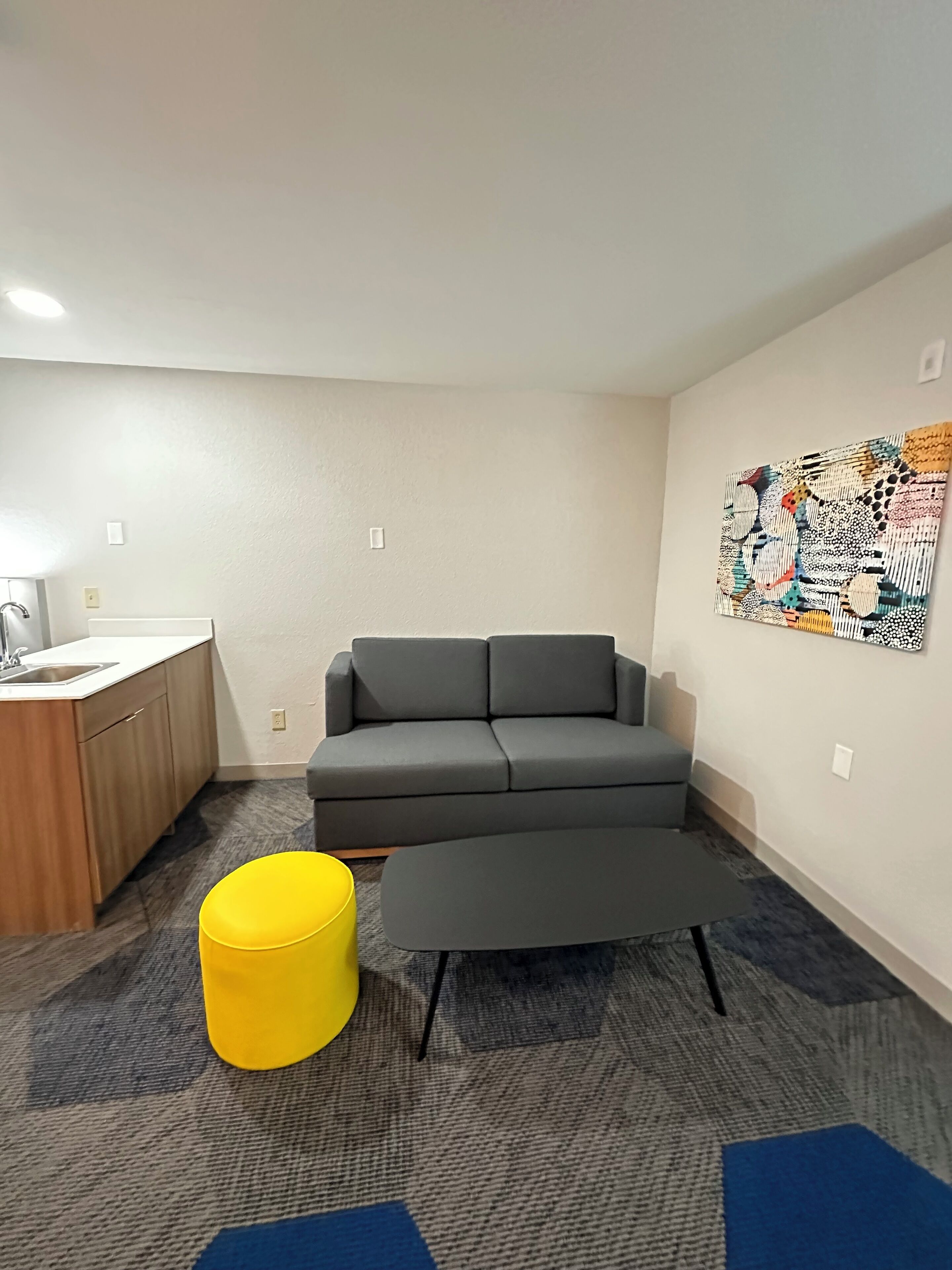 A modern living room with a gray sofa, a yellow ottoman, a gray coffee table, a wooden cabinet with a sink, and a colorful abstract painting on the wall.