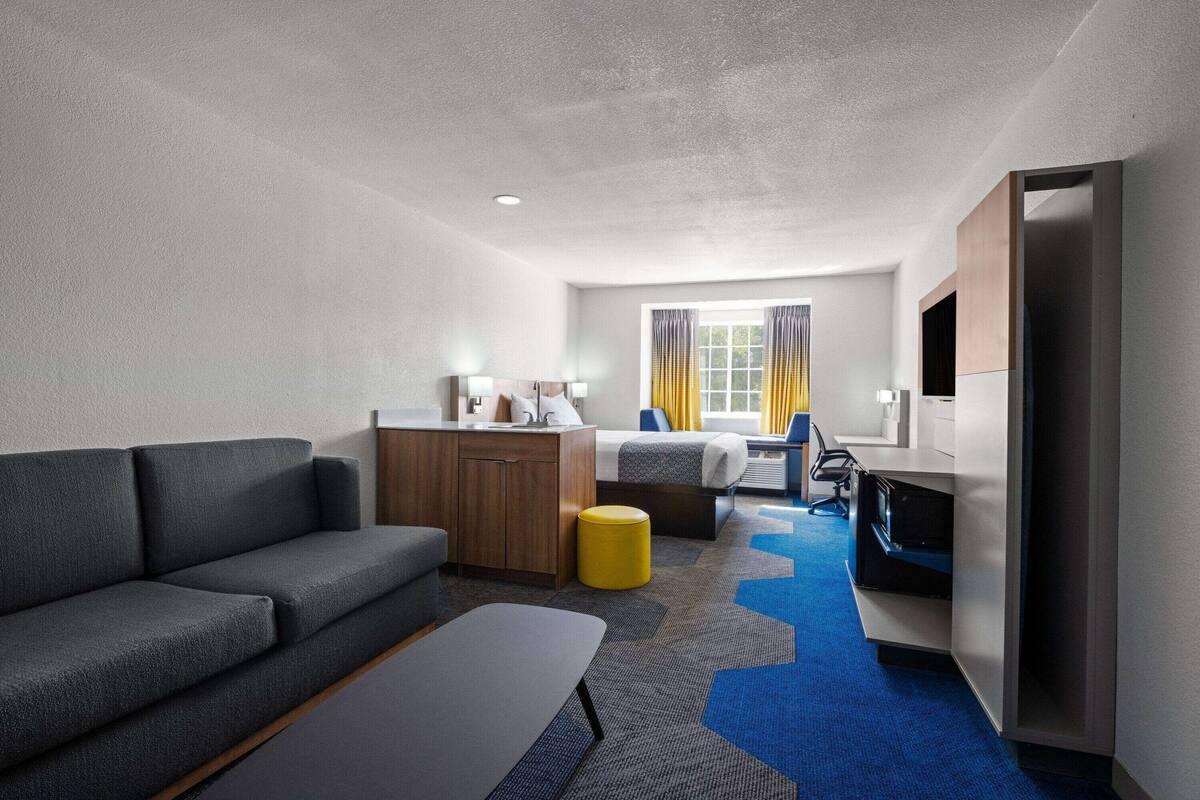 A modern hotel room with a grey sofa, a wooden desk, a bed with curtains, and a TV.